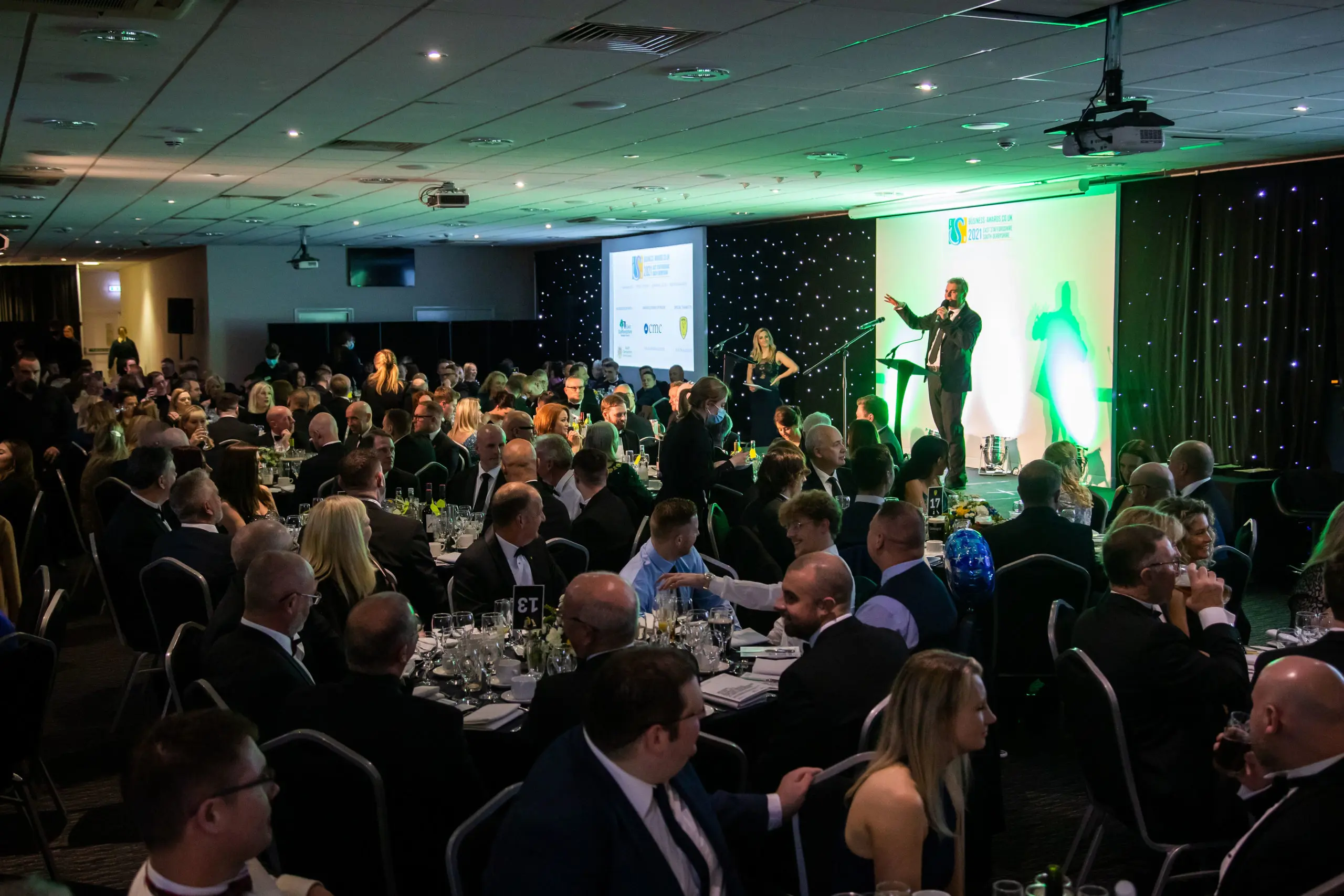 Large formal gala event with speaker on stage addressing seated dinner guests in elegant ballroom.