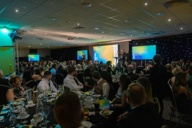 Large conference hall with hundreds of attendees seated at tables during a business presentation on stage.