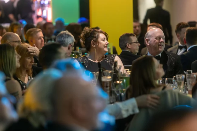 Formal dinner event with well-dressed guests seated at tables in an indoor venue with colorful backdrop.